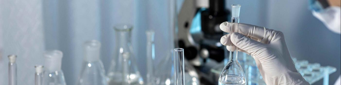 Laboratorium ze szklanymi fiolkami wypełnionymi w połowie wodą, w tle mikroskop, fragment postaci, w dłoni w rękawiczce trzyma fiolkę