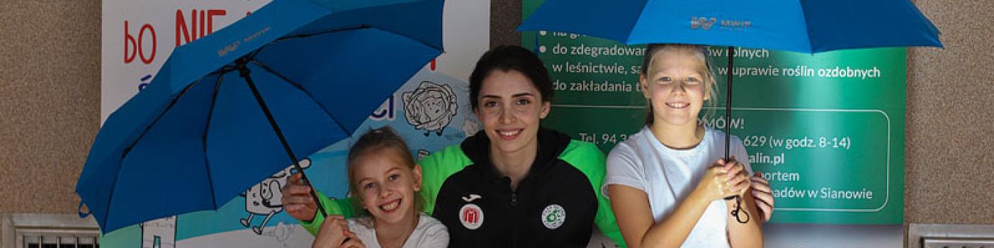 Uśmiechnięte dziewczynki przyklękają po bokach zawodniczki sportowej drużyny piłki ręcznej i trzymają w rękach parasole z logo MWiK.