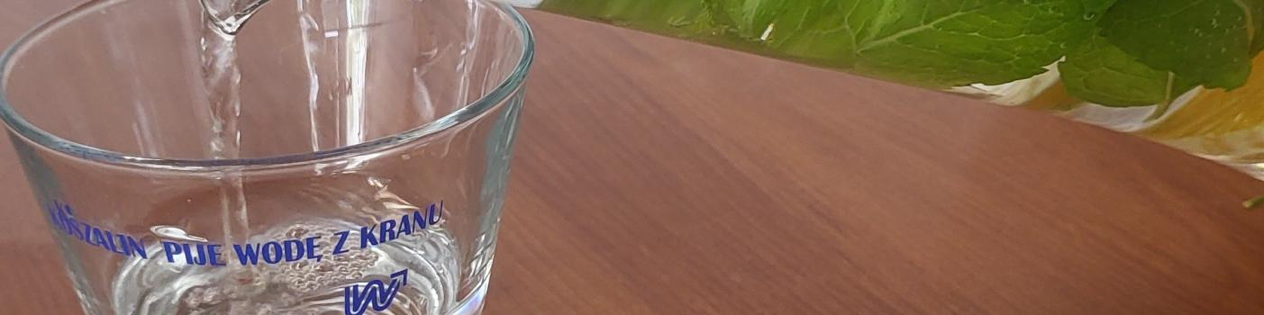 Kobieca dłoń nalewa wodę ze szklanego dzbanka do szklanki ustawionej na stole. Na dzbanku i szklance napis KOSZALIN PIJE WODĘ Z KRANU