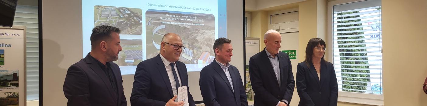 Pięcioro biznesowo ubranych osób, w tym jedna pani, stoi za stołem prezydialnym. Mężczyzna drugi od lewej prezentuje podpisaną umowę, w tle prezentacja multimedialna.