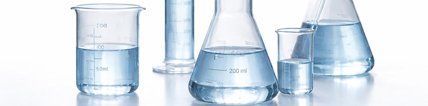 Menzurki laboratoryjne do połowy wypełnione lekko błękitną wodą stoją blisko siebie na białym blacie, wokół białe tło. W naczyniu z prawej strony znajduje się szklana pipeta. 