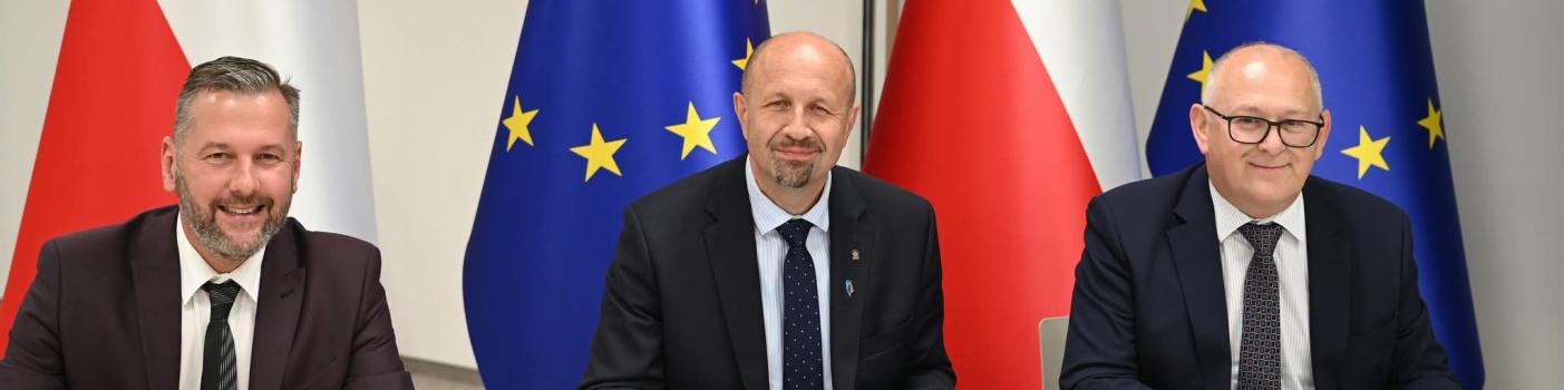 trzech biznesowo ubranych mężczyzn zasiada za stołem konferencyjnym uśmiechając się