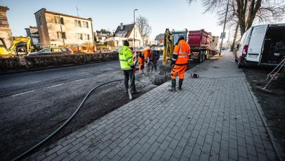 Ulica w remoncie, mężczyźni w kamizelkach odblaskowych, koparka i pojazdy specjalistyczne w tle, w dali budynki mieszkalne