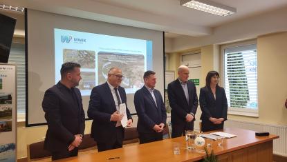 Pięcioro biznesowo ubranych osób, w tym jedna pani, stoi za stołem prezydialnym. Mężczyzna drugi od lewej prezentuje podpisaną umowę, w tle prezentacja multimedialna.