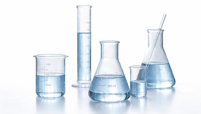 Menzurki laboratoryjne do połowy wypełnione lekko błękitną wodą stoją blisko siebie na białym blacie, wokół białe tło. W naczyniu z prawej strony znajduje się szklana pipeta. 