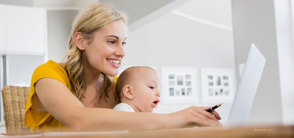 Młoda uśmiechnięta kobieta siedzi przy stole z niemowlęciem na kolanach, przed nią ekran komputera, w dłoni pióro do pisania