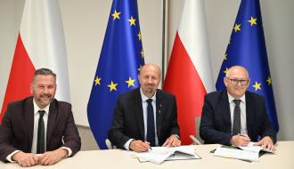 Trzech biznesowo ubranych mężczyzn zasiada za stołem konferencyjnym, uśmiechając się. Za nim flagi Polki i Unii Europejskiej.