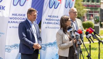 Dwóch biznesowo ubranych mężczyzn i jedna kobieta stojąca pomiędzy nimi w środku udzielają wywiadu przed mikrofonami. Za nimi flagi flagi z logo MWiK.