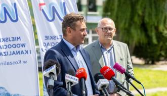 Dwóch biznesowo ubranych mężczyzn przed mikrofonami udziela wywiadu. Za nimi flagi flagi z logo MWiK.